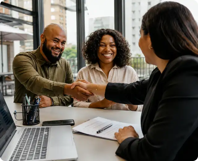 A smiling couple shaking hands with a professional advisor across a desk with a laptop and paperwork.